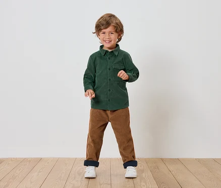 Un garçon debout souriant porte une chemise verte, un pantalon marron et des chaussures blanches.