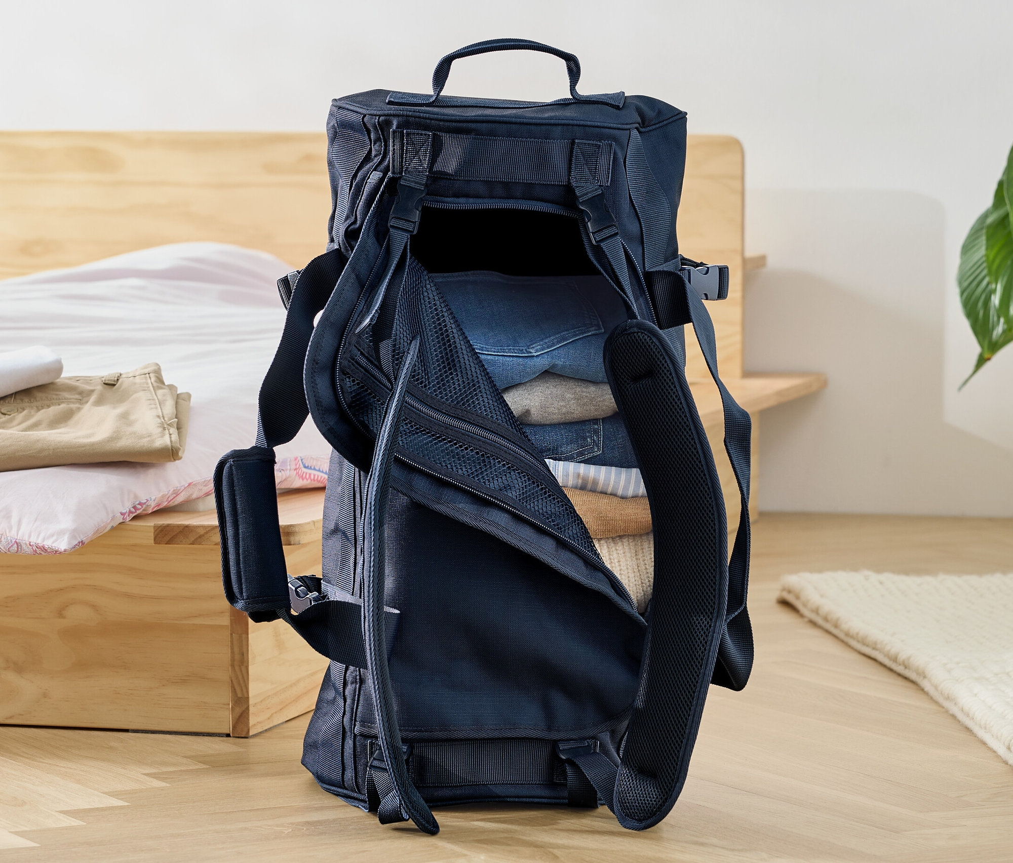 Un sac de voyage et sac à dos ouvert rempli de vêtements posé sur le sol à côté d'un lit.