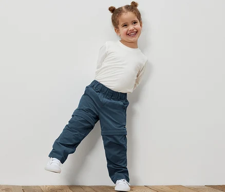 Une petite fille souriante appuyée contre un mur blanc porte un pantalon fonctionnel pour tout-petit et une chemise blanche.
