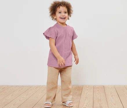 Fillette souriante posant en blouse en mousseline, pantalon et sandales.