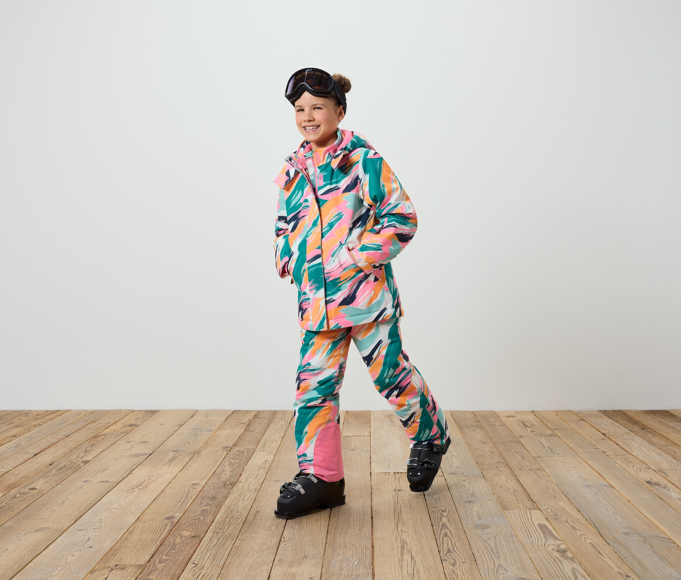 Une fillette souriante pose en combinaison de ski à rayures colorées et chaussures de ski.