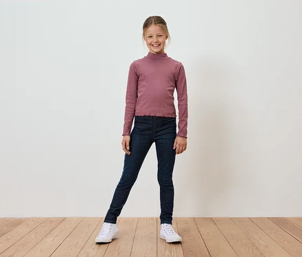 Une fillette souriante portant un haut rose à manches longues, un jegging bleu marine et des baskets blanches se tient sur un plancher de bois devant un mur blanc.