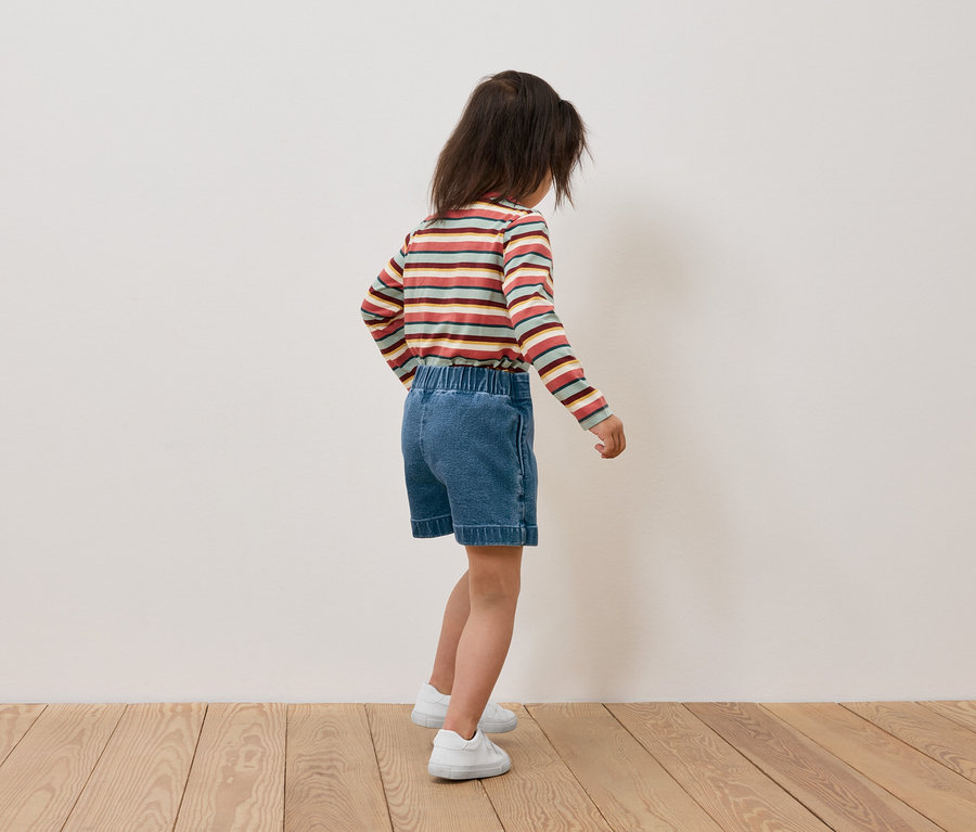 Fillette portant un t-shirt rayé à manches longues et un short en jean, dos à la caméra.
