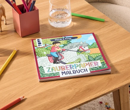 Sur une table se trouvent un livre de coloriage Zauberpapier Malbuch, des crayons de couleur, un verre d'eau et deux figurines de chevaux.