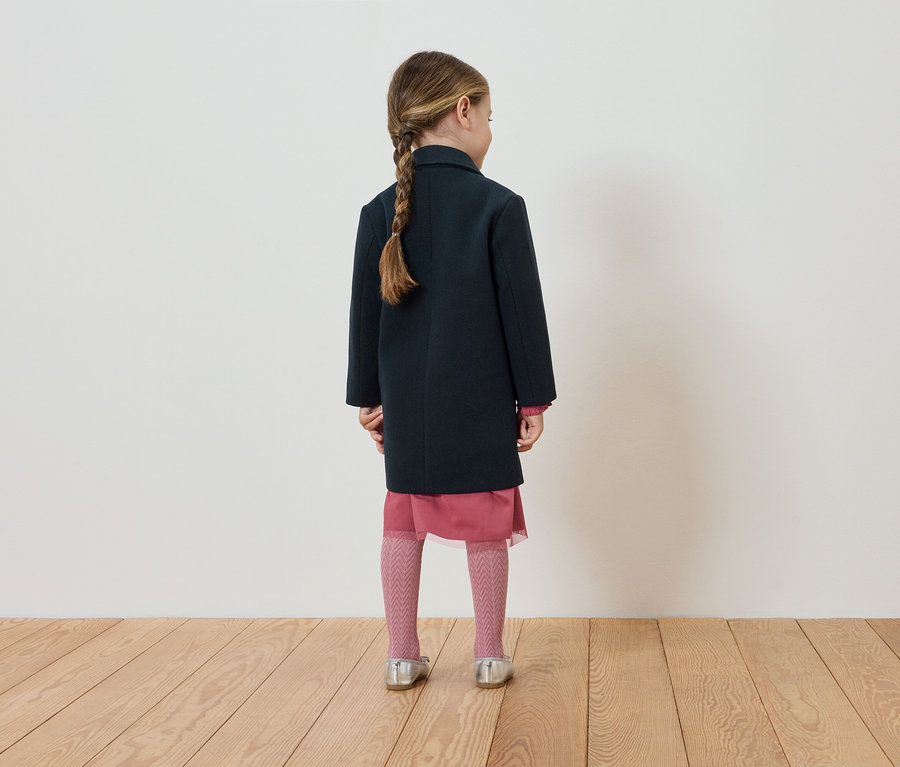Une fillette, vue de dos, porte un manteau bleu marine, une robe rose, des collants et des chaussures argentées.