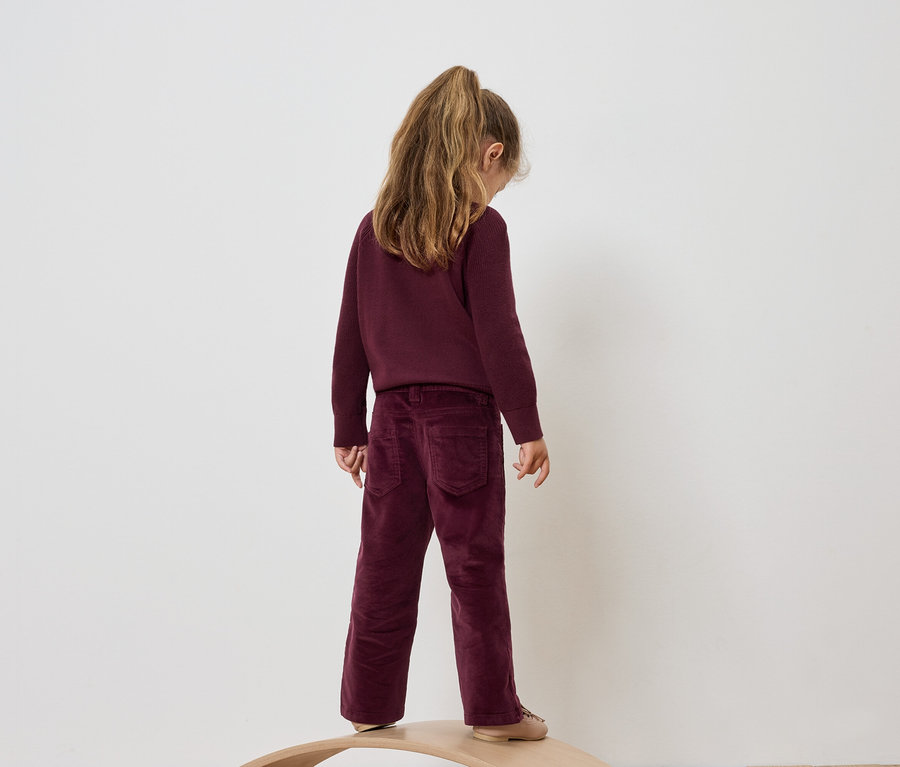 Une fille portant un pantalon et un pull en velours bordeaux, se tenant dos tourné et regardant vers le bas.