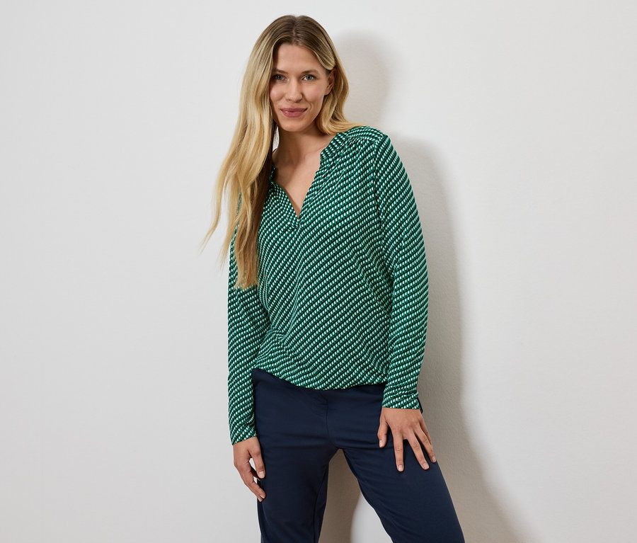 Une femme blonde pose en chemisier vert rayé et pantalon bleu marine.