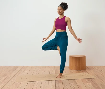 Une femme en haut rose et pantalon bleu se tient en position de l'arbre sur un tapis de yoga.