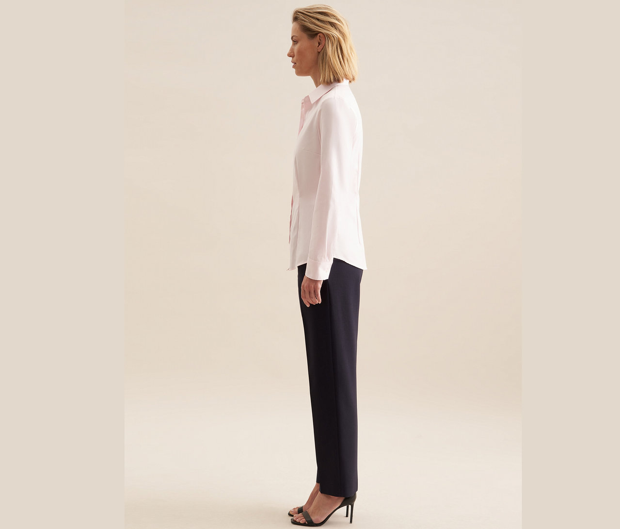 Une femme se tient de profil et porte un chemisier en tulle Seidensticker rose, un pantalon bleu marine et des sandales à talons noires.