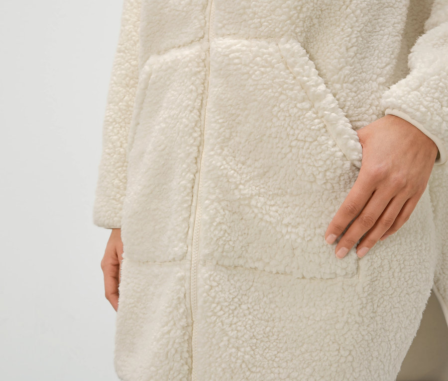 Gros plan d'un peignoir crème en fausse fourrure pour femme avec fermeture éclair et poches, main dans la poche.