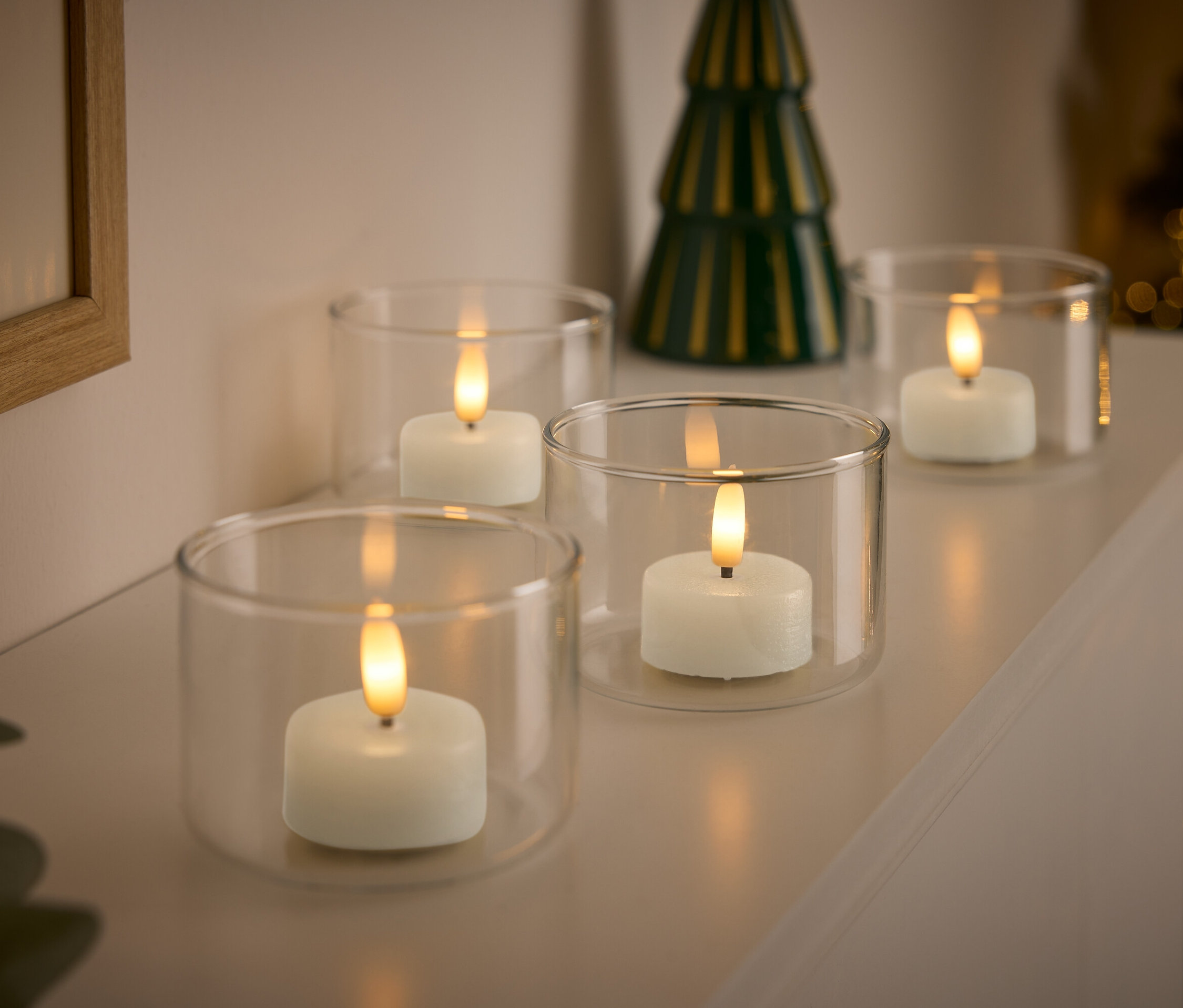 Quatre bougies blanches dans des bougeoirs en verre transparent avec un petit sapin de Noël vert en arrière-plan.