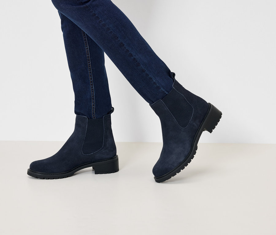 Jambes en jean bleu marine et bottines Chelsea en daim bleu marine à talons bas marchant sur une surface blanche.