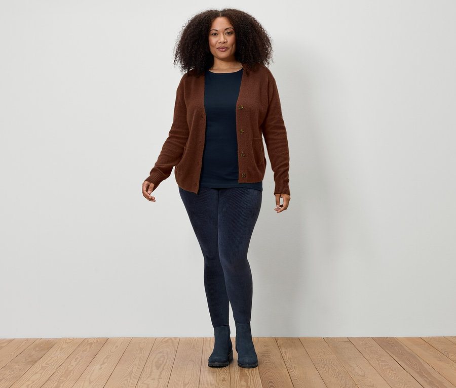 Une femme aux cheveux bouclés bruns se tient devant un mur blanc et porte un cardigan marron, un t-shirt bleu marine, un legging bleu marine et des bottes bleu marine.