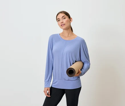 Femme tenant un tapis de yoga enroulé, portant un haut bleu à manches longues et un legging noir.