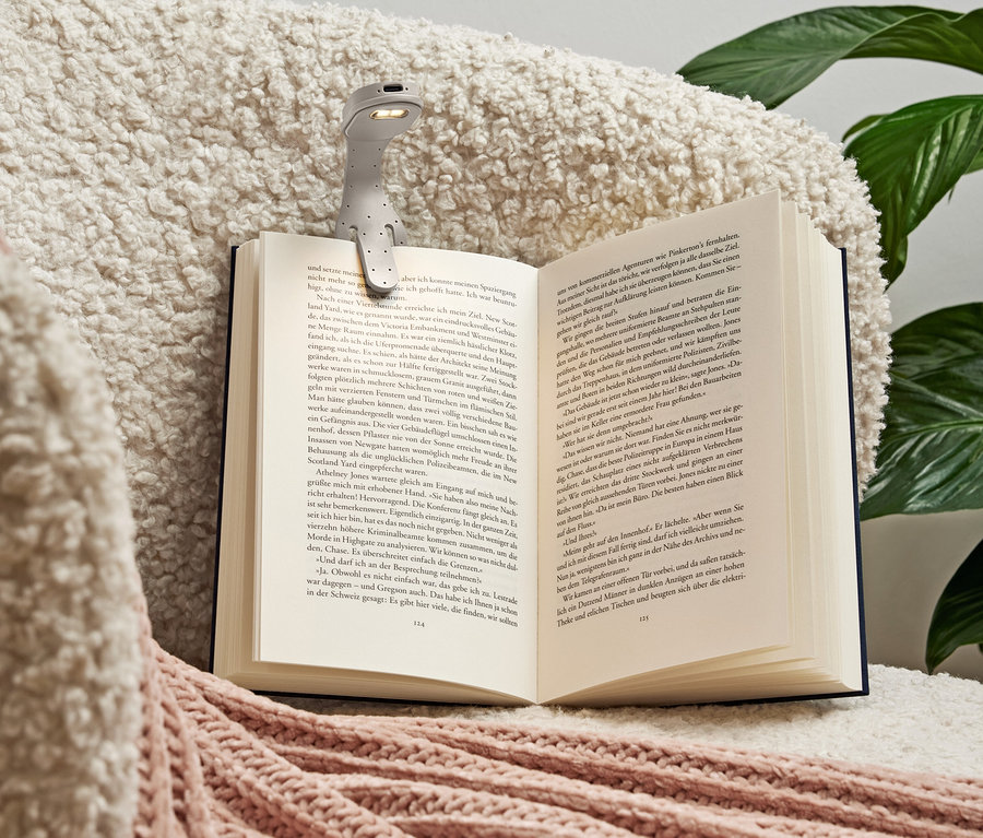 Une lampe de lecture grise est fixée à un livre ouvert posé sur une couverture douce de couleur crème avec des accents roses.