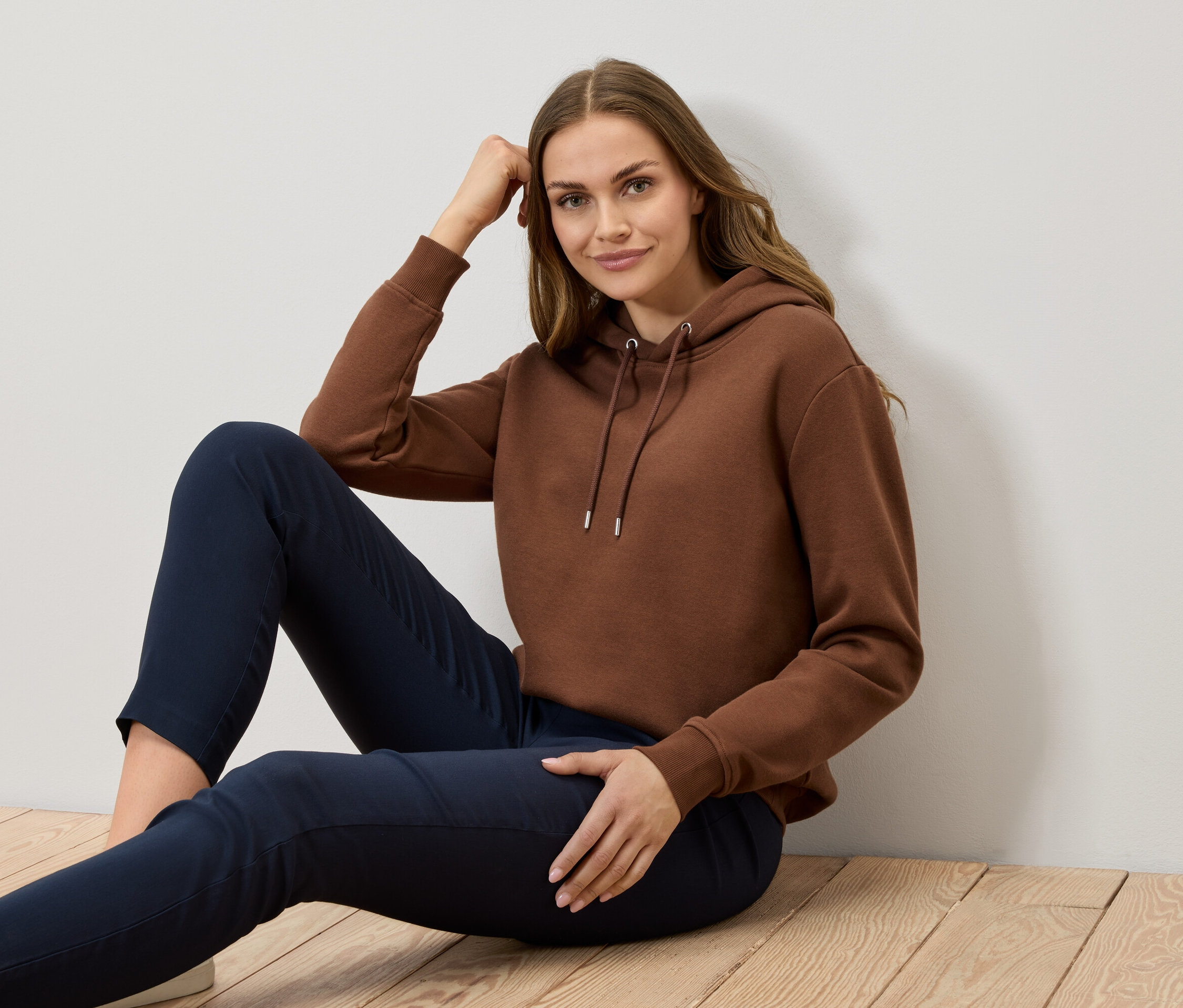 Une femme est assise sur un plancher en bois, portant un sweat à capuche marron et un pantalon bleu marine.