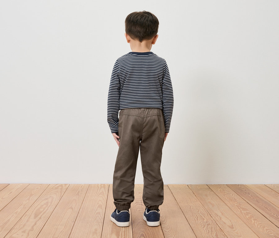 Un garçon se tient dos à la caméra, portant un t-shirt rayé, un pantalon marron et des chaussures bleues.