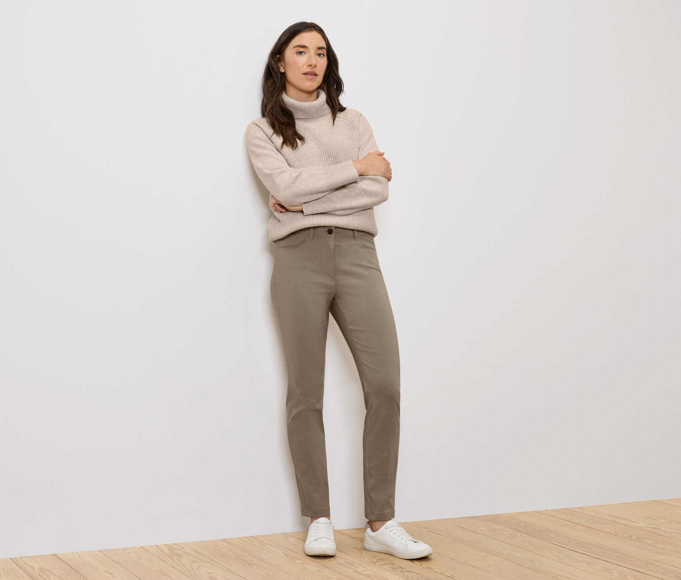 Une femme se tient debout, les bras croisés, portant un pull à col roulé beige, un pantalon marron et des baskets blanches.