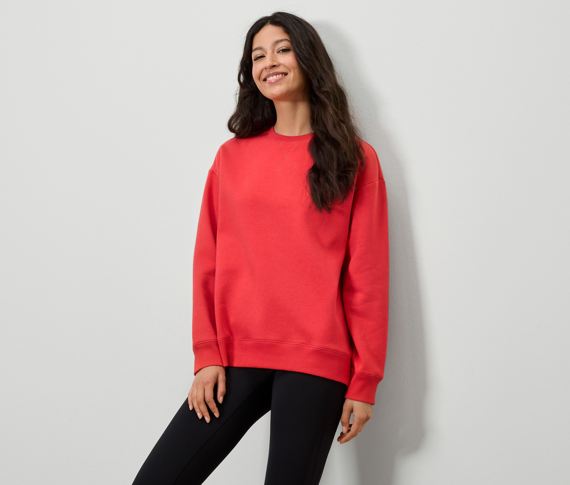 Une femme portant un sweat-shirt rouge et des leggings noirs pose devant un mur blanc.