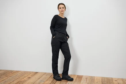 Femme debout, mains dans les poches, portant un pantalon noir et des bottes noires.