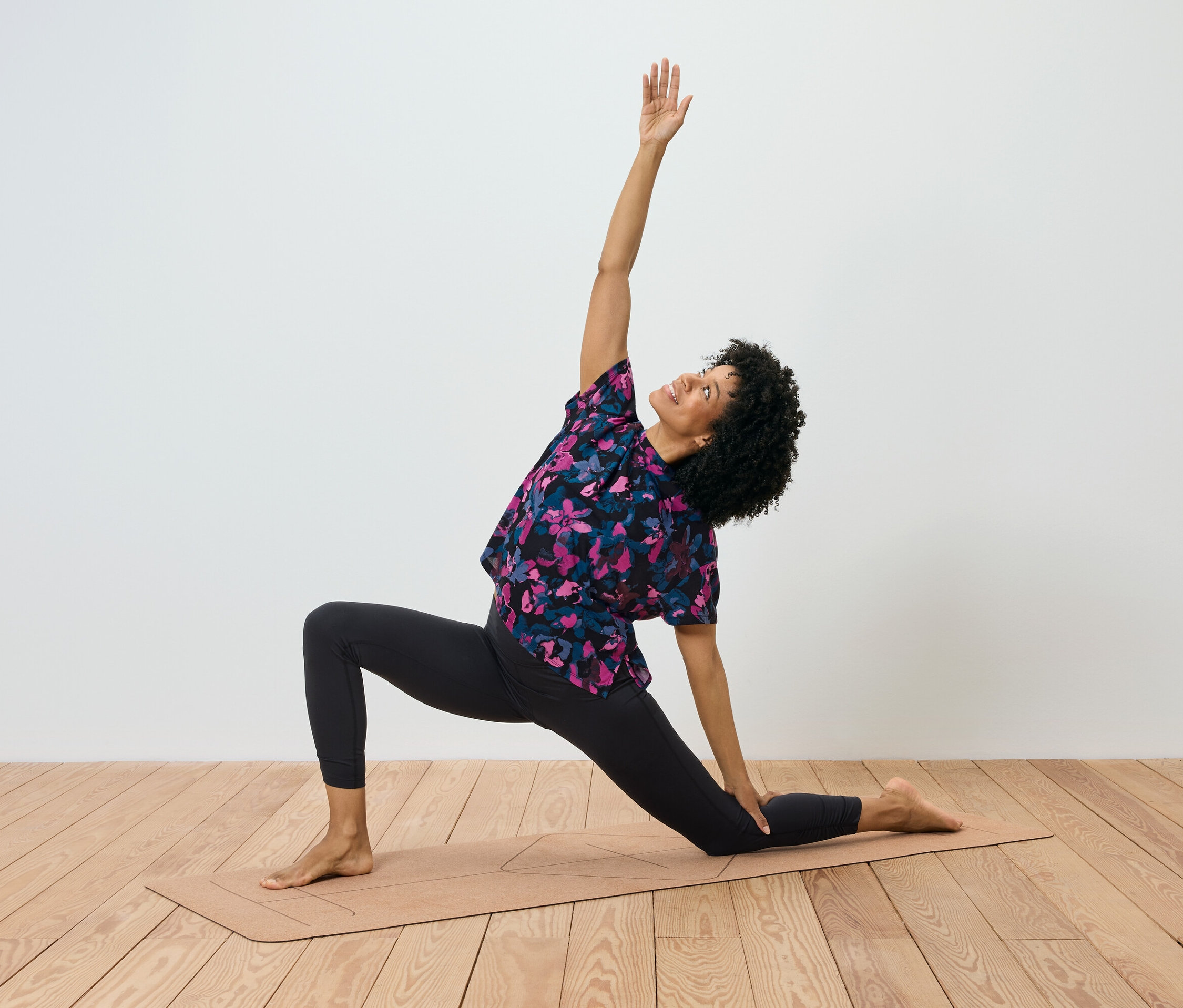 Une femme aux cheveux bouclés fait un exercice sur un tapis de yoga, le bras droit est levé et le gauche touche le tapis.