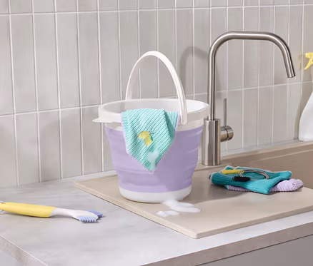 Un seau pliable violet avec un chiffon brodé de citron est posé sur le comptoir à côté d'un évier, où se trouvent une brosse jaune et des chiffons.