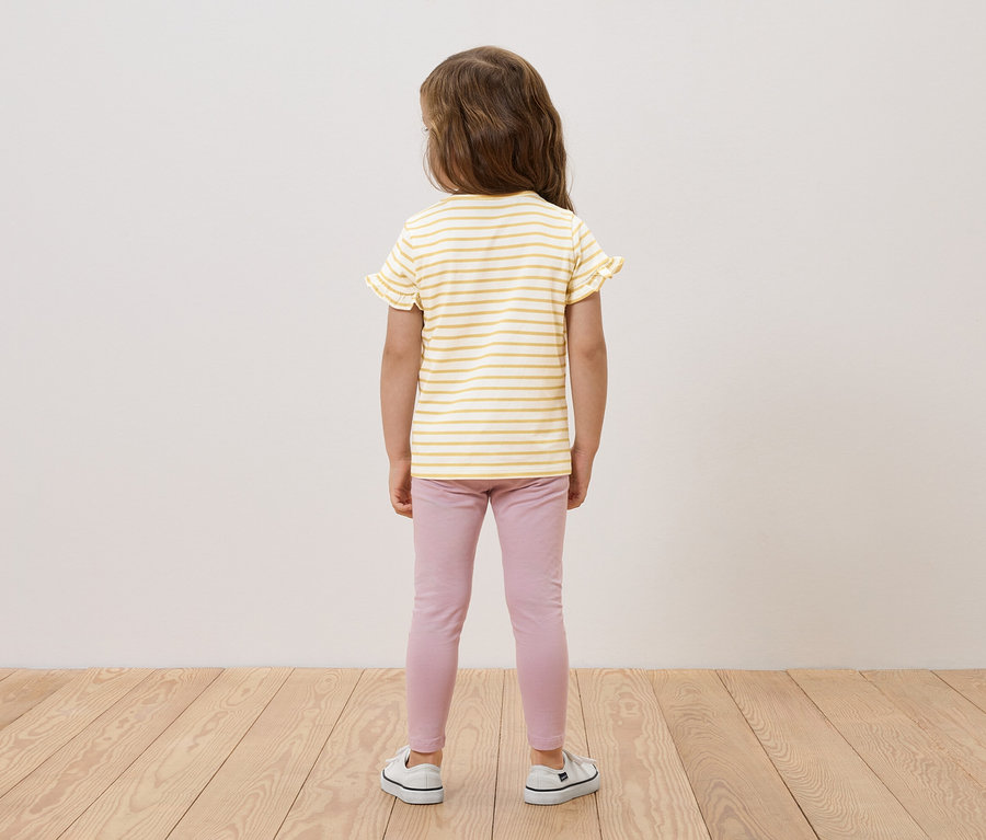 Une fillette, vue de dos, porte un haut rayé jaune et blanc à volants, un legging rose et des baskets blanches.