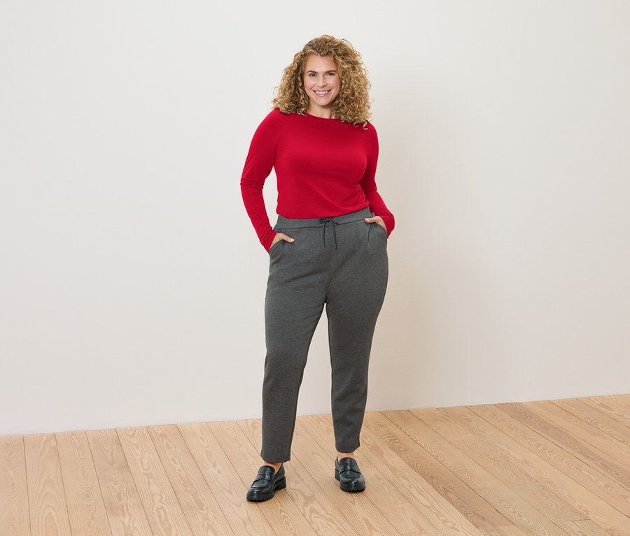 Une femme aux cheveux bouclés se tient devant un mur blanc, portant un haut rouge à manches longues et un pantalon de jogging gris.