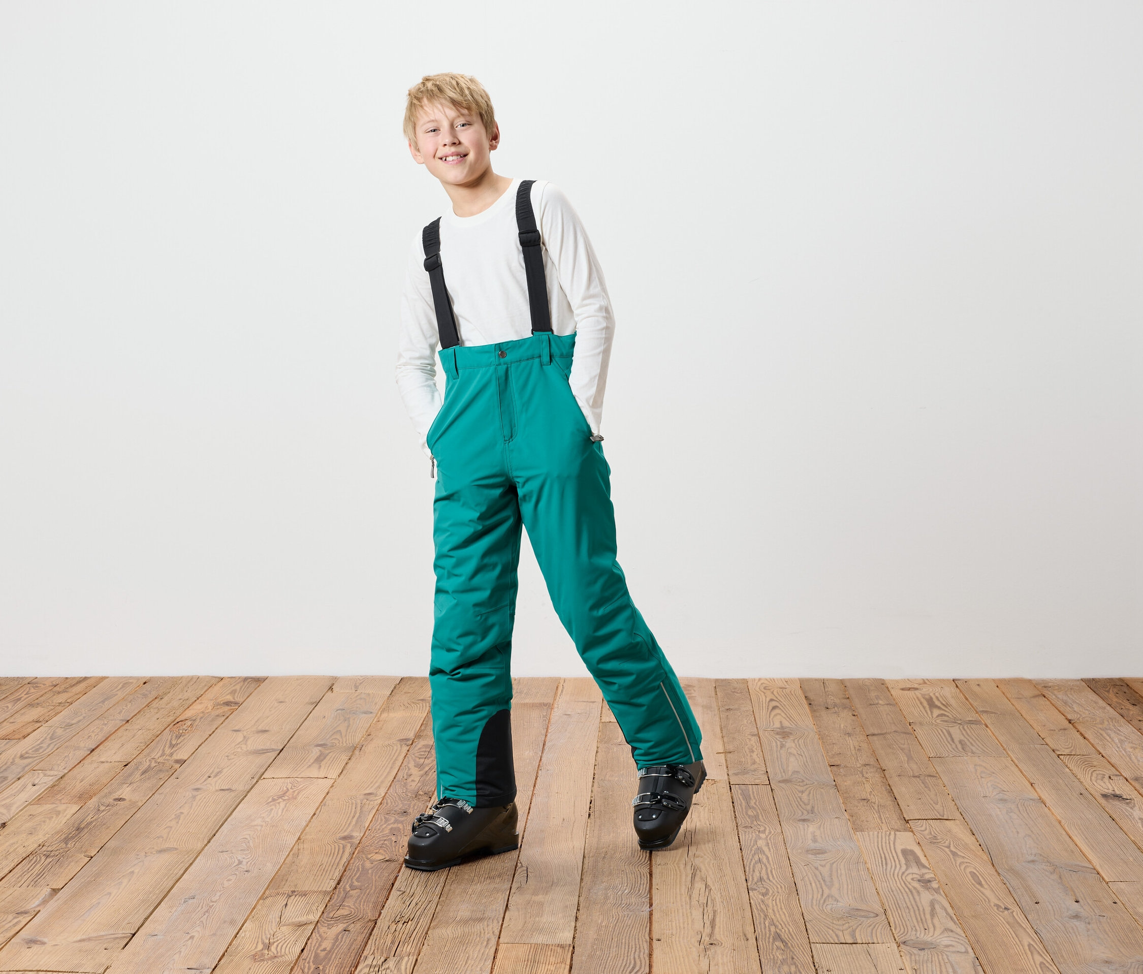 Un garçon portant un pantalon de ski vert avec des bretelles noires et des chaussures de ski se tient sur un plancher en bois devant un mur blanc.