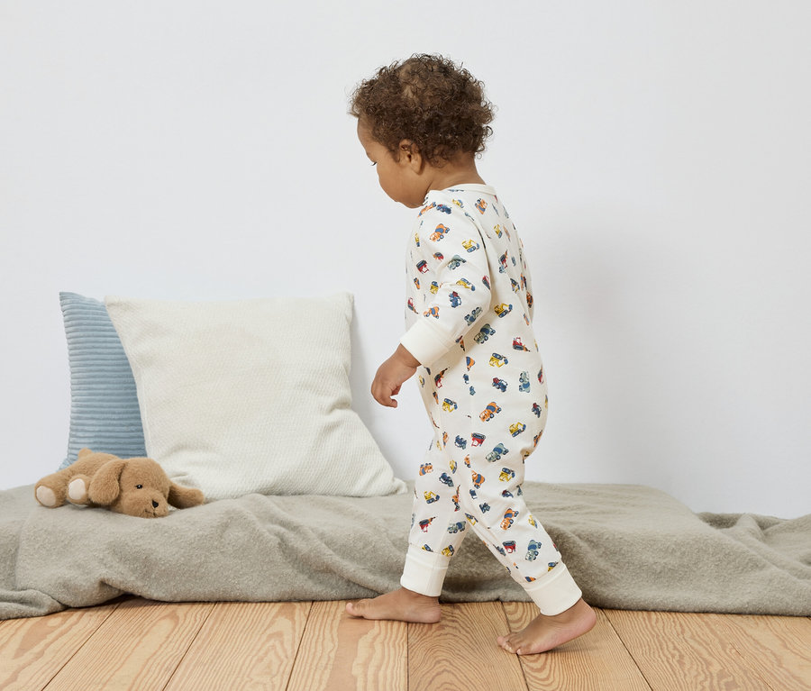 Un jeune enfant aux cheveux bouclés marche pieds nus sur un plancher en bois. Il porte un pyjama crème avec des motifs de voitures colorées. À l'arrière-plan, on peut voir des coussins et un chien en peluche.