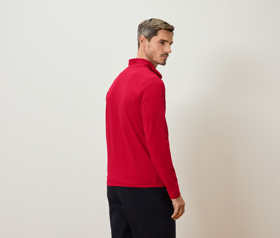 Homme de profil, portant un t-shirt rouge à manches longues et un pantalon foncé sur fond clair.
