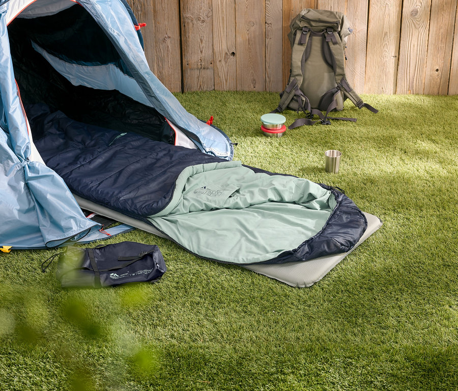 Sur l'herbe se trouvent une tente ouverte, un sac de couchage sur un matelas de sol, un sac à dos, une boîte et une tasse.