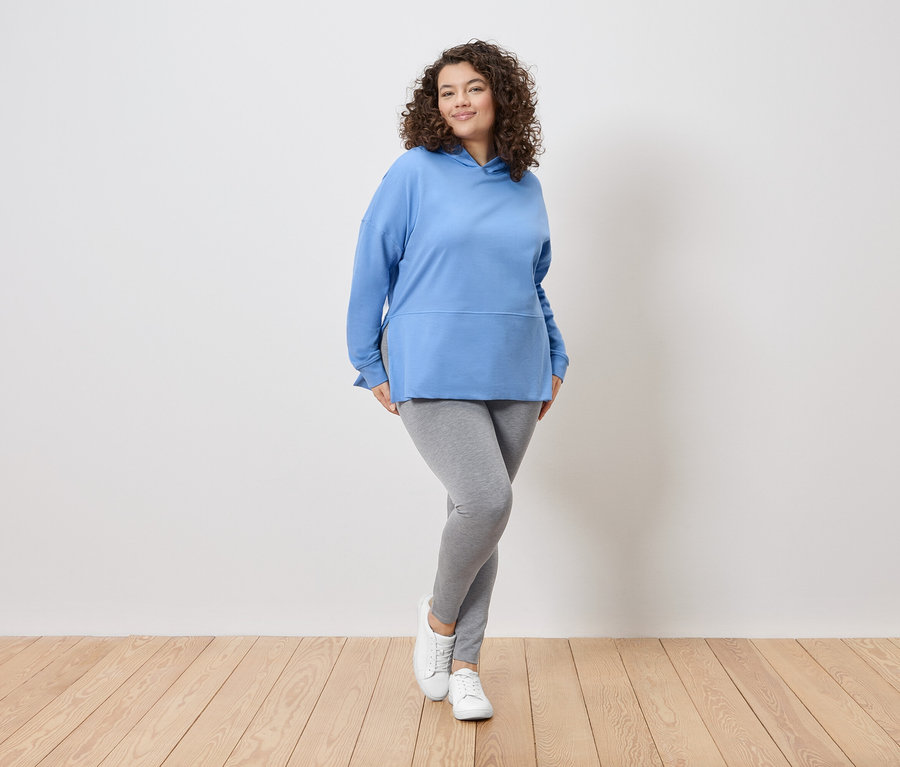 Une femme aux cheveux bouclés se tient devant un mur blanc, portant un sweat à capuche bleu, un legging gris et des baskets blanches.