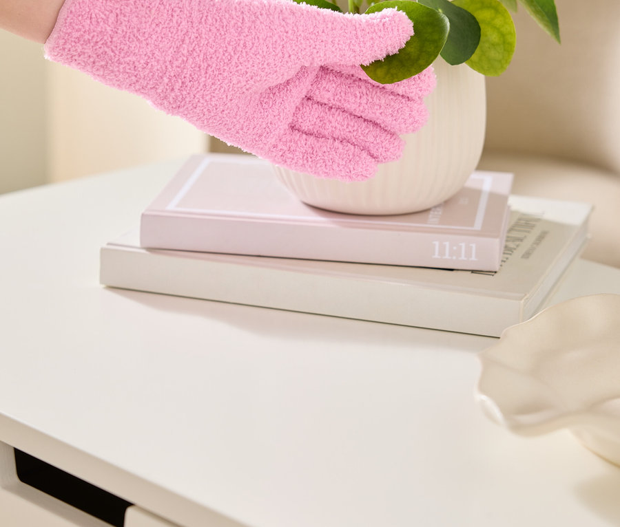 Une main gantée d'un gant rose et duveteux touche les feuilles d'une plante d'intérieur dans un pot blanc, posé sur deux livres sur une table blanche.