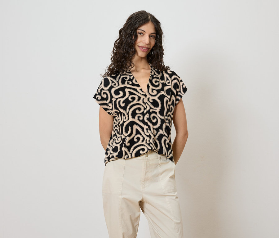Une femme aux cheveux bouclés porte un chemisier à motifs noir et beige et un pantalon beige, posant devant un mur blanc.