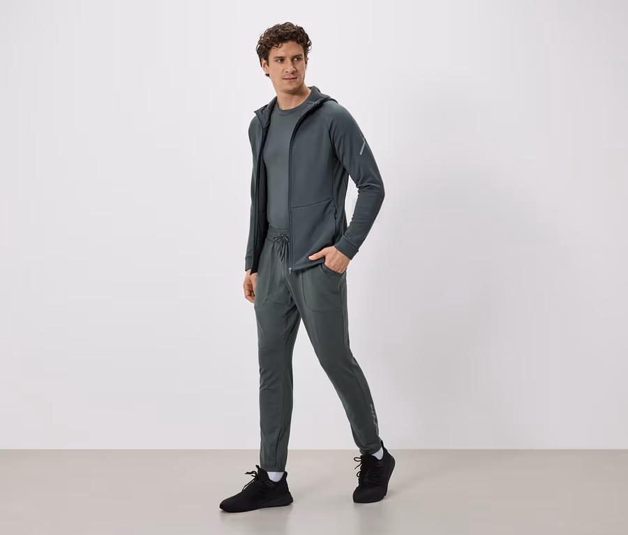 Homme en survêtement vert olive avec veste à capuche et pantalon à cordon de serrage, et baskets noires.