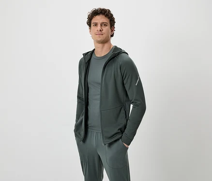 Un homme aux cheveux bouclés porte un survêtement vert foncé composé d'un sweat à capuche, d'un t-shirt et d'un pantalon de jogging, sur un fond neutre.