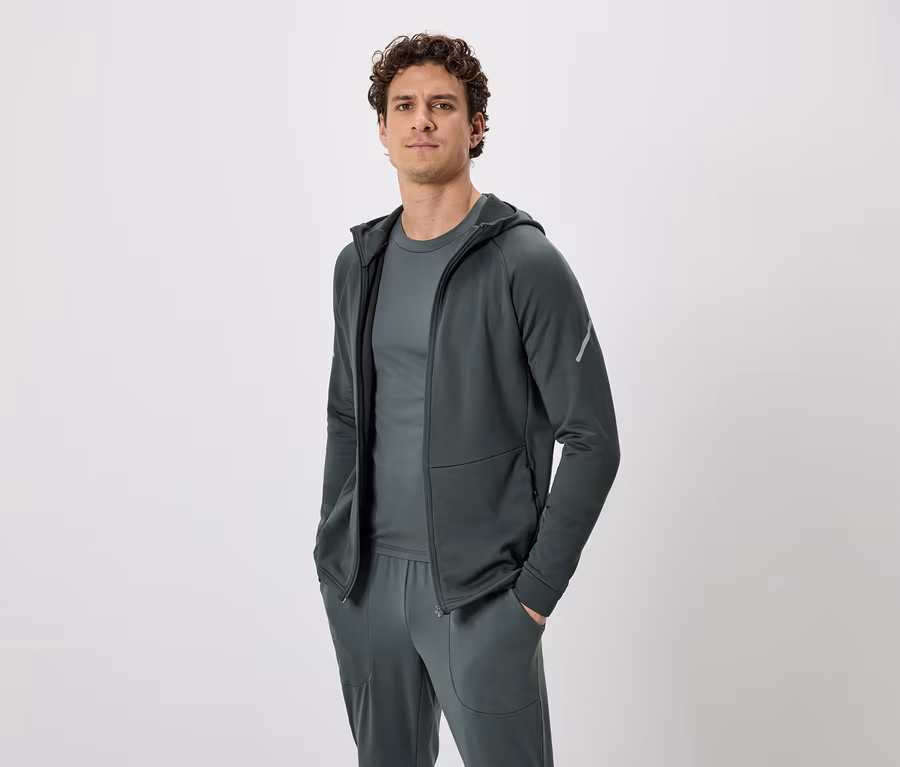Un homme aux cheveux bouclés porte un survêtement vert foncé composé d'un sweat à capuche, d'un t-shirt et d'un pantalon de jogging, sur un fond neutre.