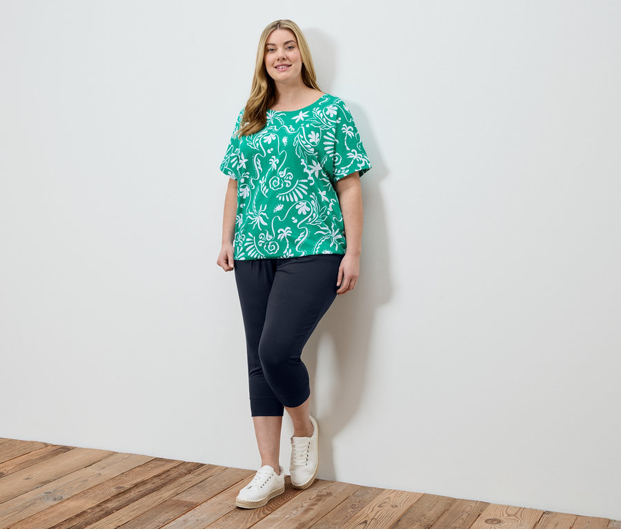 Une femme portant un haut vert à motifs blancs, un pantalon bleu marine et des chaussures blanches se tient devant un mur blanc.