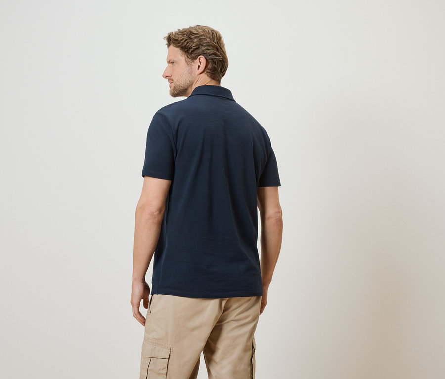 Homme vu de dos portant un polo bleu marine et un pantalon cargo beige sur fond blanc.