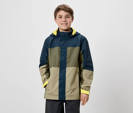 Un garçon souriant se tient devant un fond blanc, portant une veste à blocs de couleurs avec une doublure jaune.