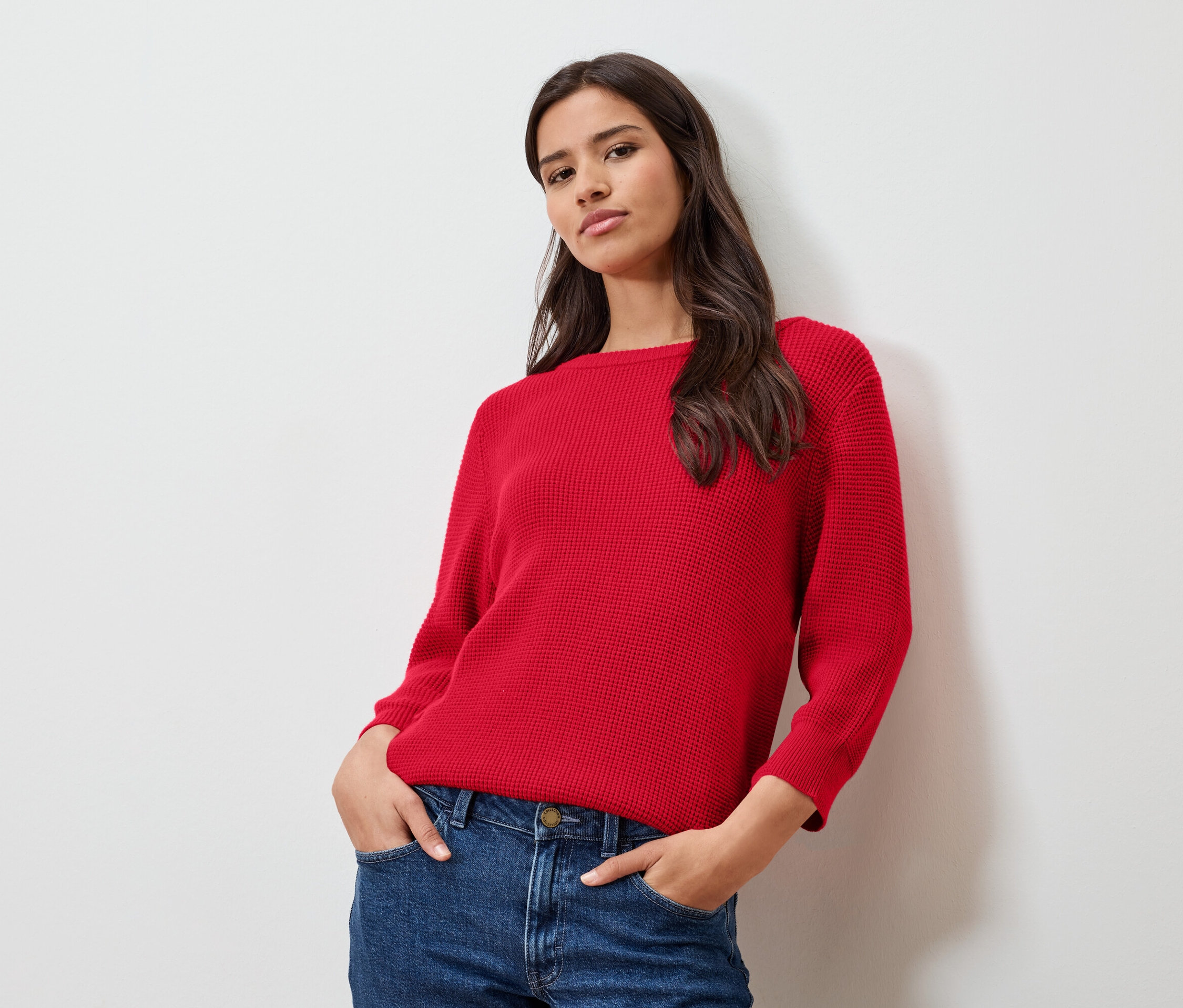 Femme portant un pull en maille fine rouge et un jean posant devant un mur blanc.