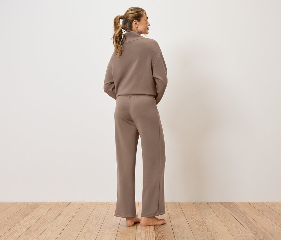 Une femme se tient dos à la caméra, vêtue d'un tailleur pantalon marron.