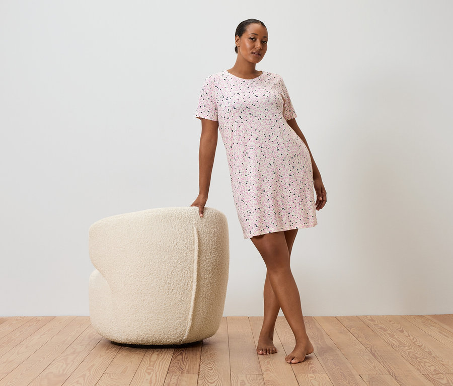 Une femme s'appuie sur un fauteuil crème, elle porte une robe rose à pois à manches courtes.