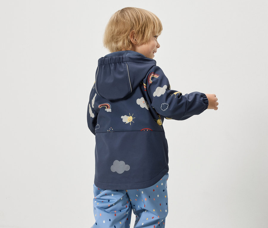 Un enfant porte un manteau de pluie bleu marine à capuche imprimé de nuages, de soleils et d'arcs-en-ciel, ainsi qu'un pantalon de pluie bleu à pois colorés.
