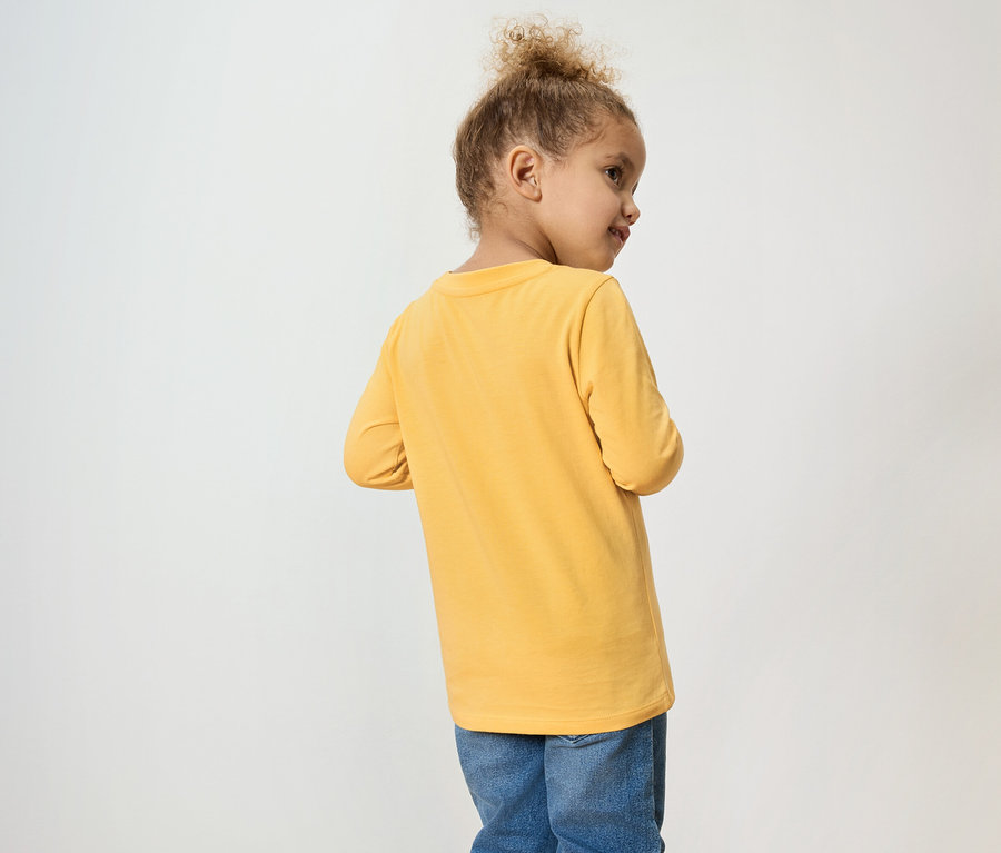 Une jeune fille aux cheveux bouclés porte un t-shirt jaune à manches longues et un jean.