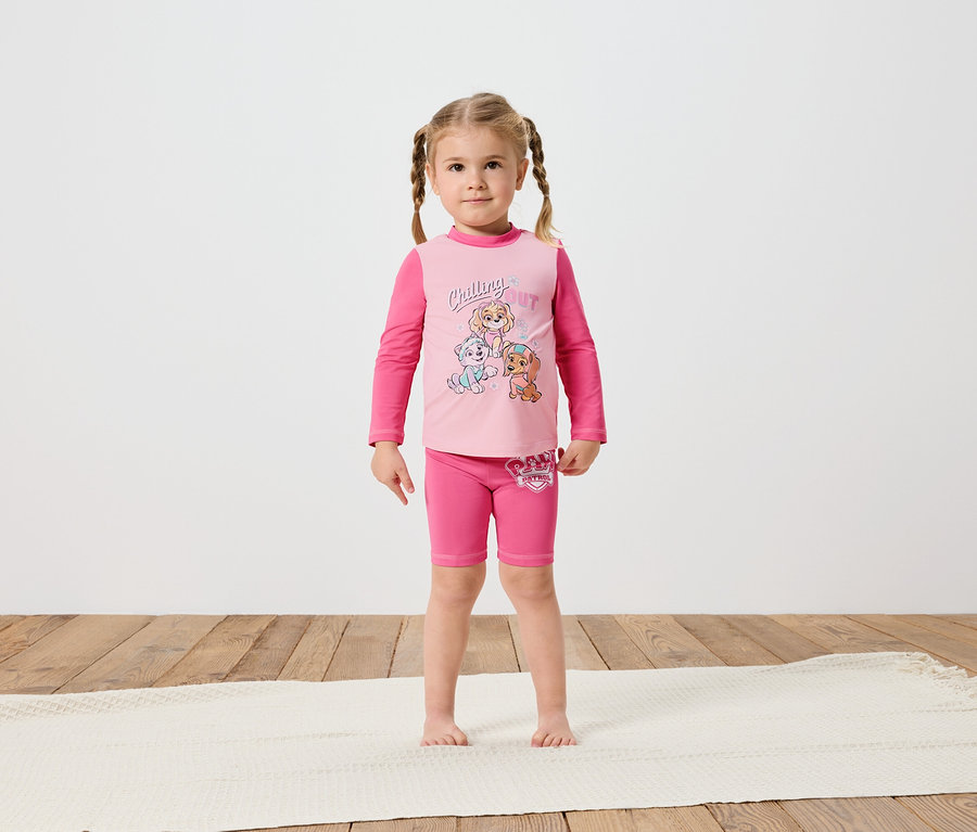 Une petite fille avec des tresses se tient pieds nus sur un tapis couleur crème, portant un T-shirt à manches longues rose et un short Pat' Patrouille.