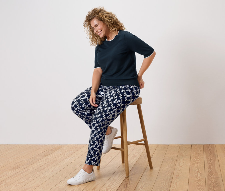Une femme aux cheveux bouclés est assise sur un tabouret, portant un haut bleu marine et un pantalon à motifs.