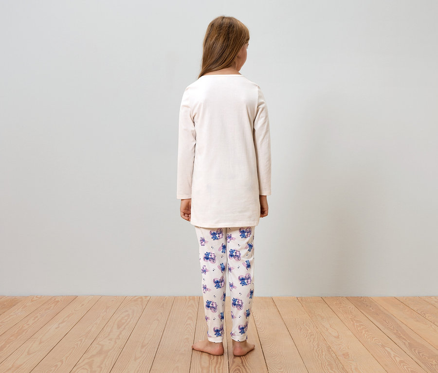 Fillette vue de dos, portant un pyjama beige avec des motifs Stitch, debout sur un plancher en bois devant un mur gris.