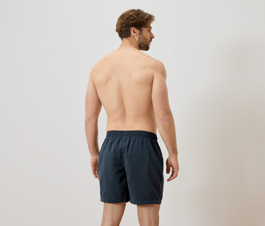 Homme vu de dos, torse nu et portant un short de bain foncé sur fond blanc.